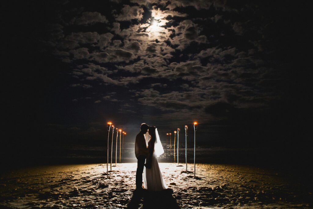 "Verlies jezelf in de magie van een bruiloft bij maanlicht, vastgelegd in één adembenemende foto. Geniet van het romantische schouwspel terwijl een bruidspaar zich omringd door fakkels bevindt op het strand, klaar voor een intieme ceremonie onder de sterrenhemel. Laat deze beeldschone foto je meenemen naar een wereld van liefde, avontuur en betovering."